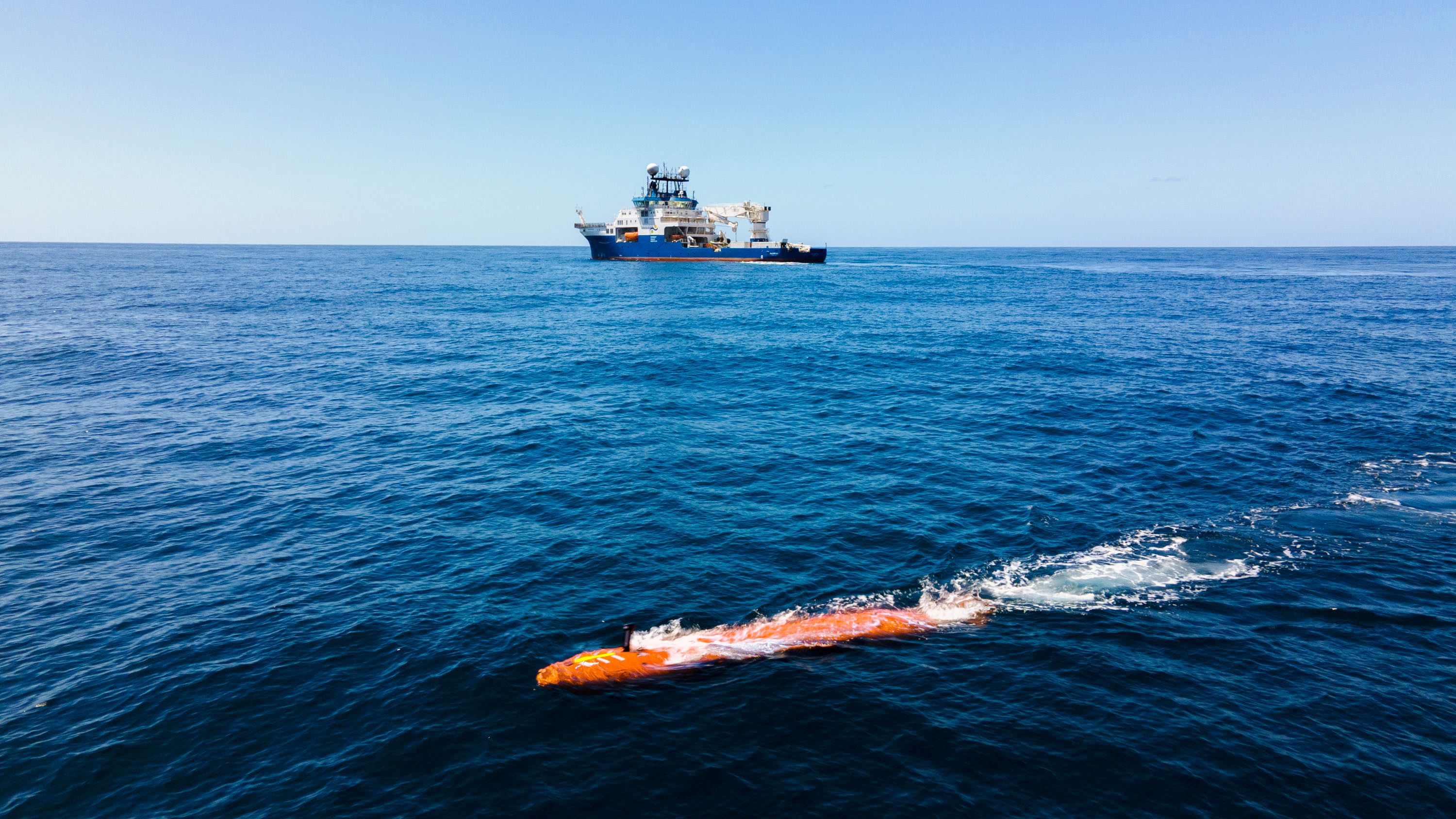 An orange, torpedo-shaped autonomous underwater vehicle rests at the blue ocean's surface, while R/V Falkor (too) looms against the horizon.