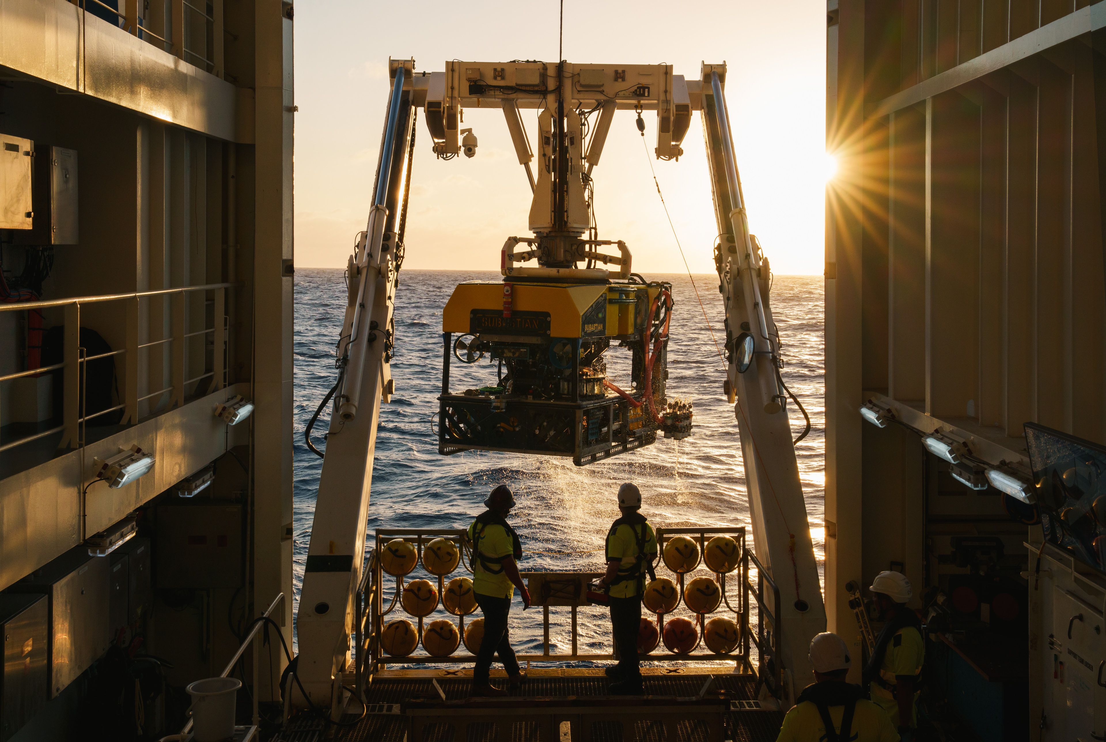 ROV Subastian being recovered with two ROV pilots operating the winch with a sun starburst peaking over the ROV.