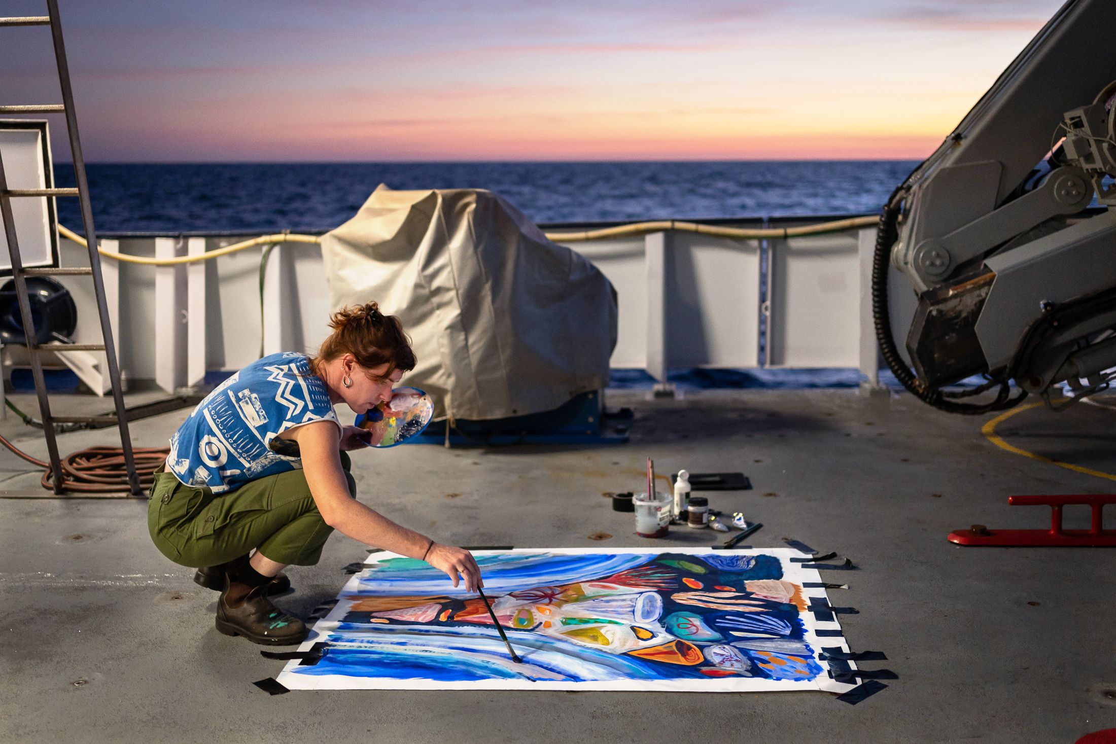person crouched down painting on a canvas on a ship deck