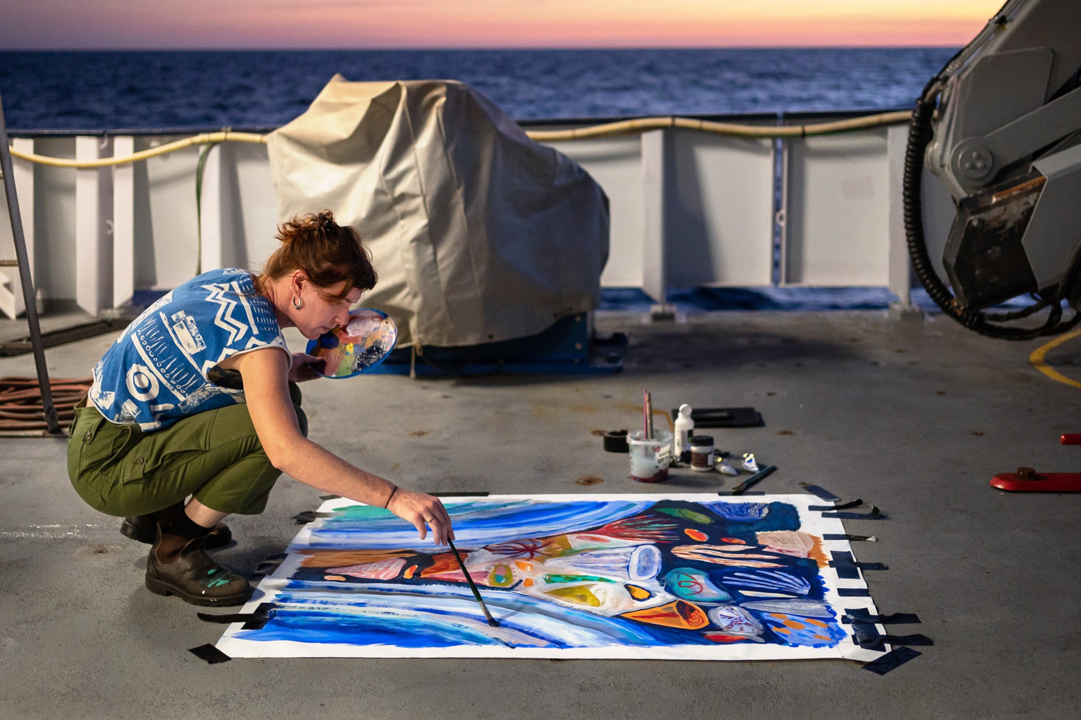 person crouched down painting on a canvas on a ship deck