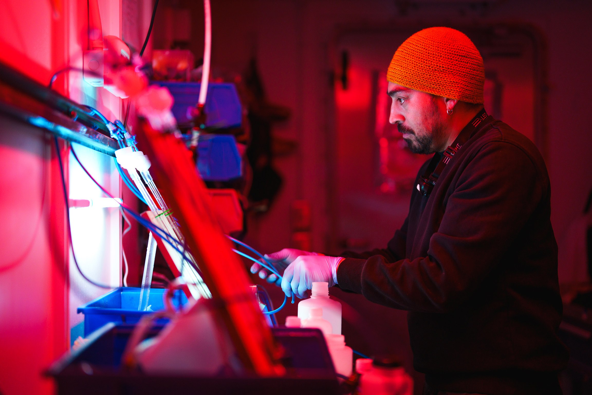 In red lighting a man with a beanie is working on some wires.