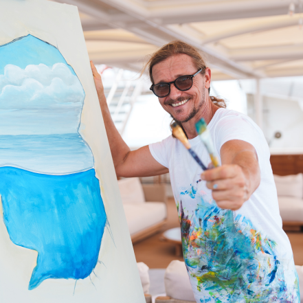 person holds paintbrushes towards camera and smiles next to canvas