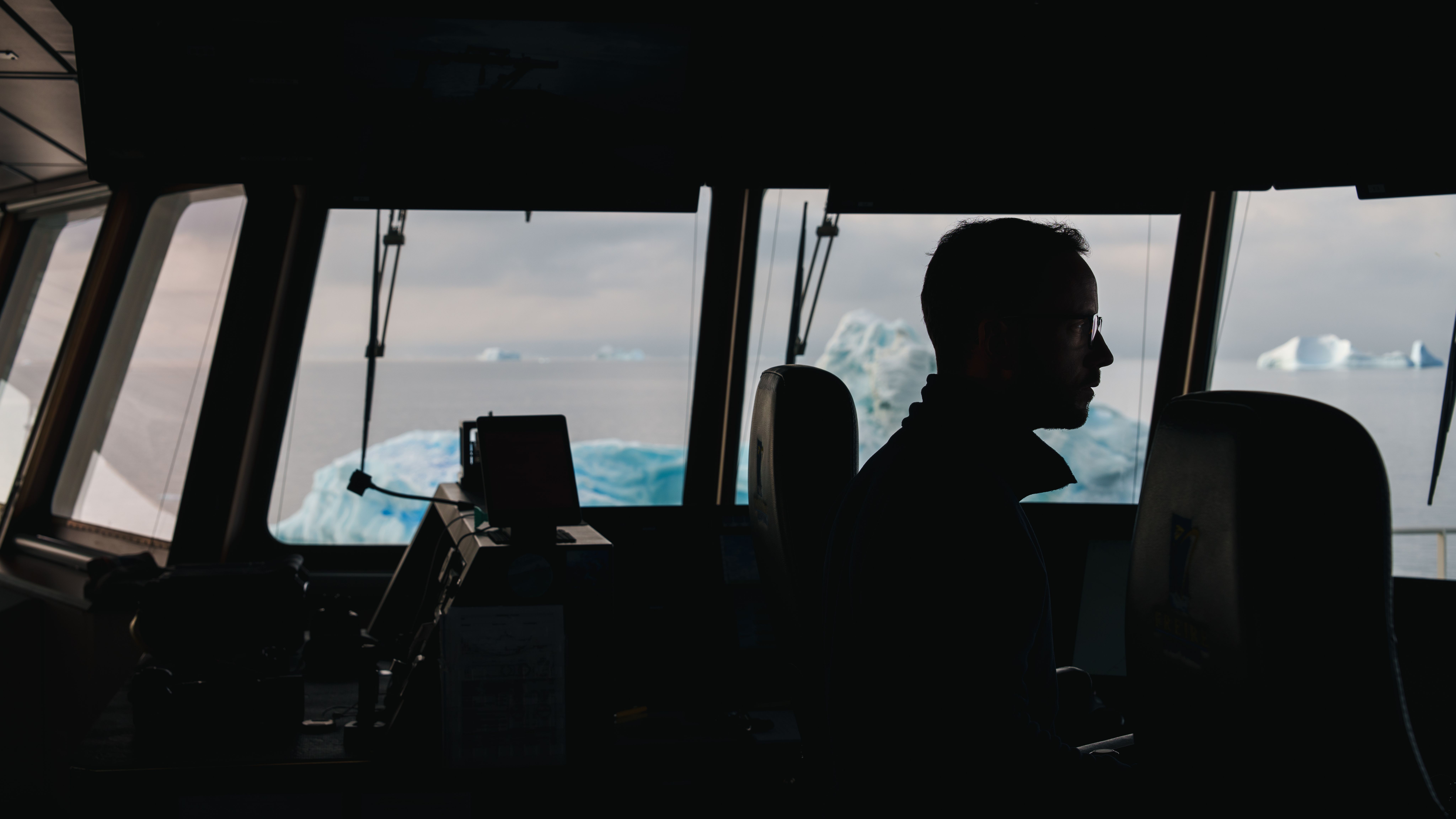 Silhouette  of a man at a window, an iceberg floats on ocean behind. 