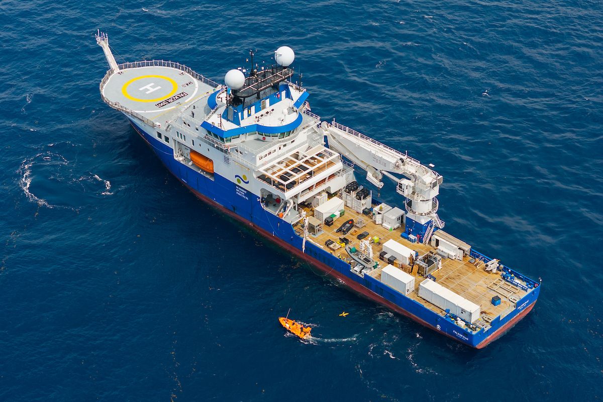 High-angle aerial photograph of the research vessel Falkor (too) at sea during glider recovery operations, with a small orange workboat deployed nearby.