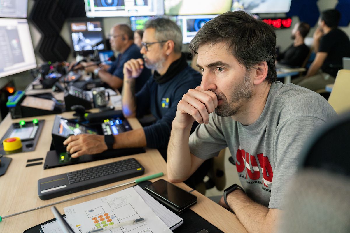 Man in a grey t-shirt resting his chin on his hand while intently watching screens alongside colleagues in a shipboard mission control room.