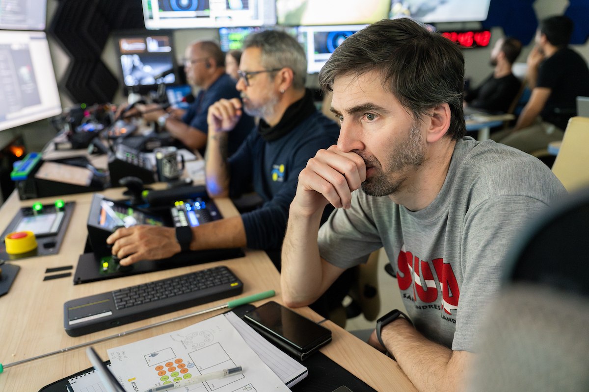 Man in a grey t-shirt resting his chin on his hand while intently watching screens alongside colleagues in a shipboard mission control room.