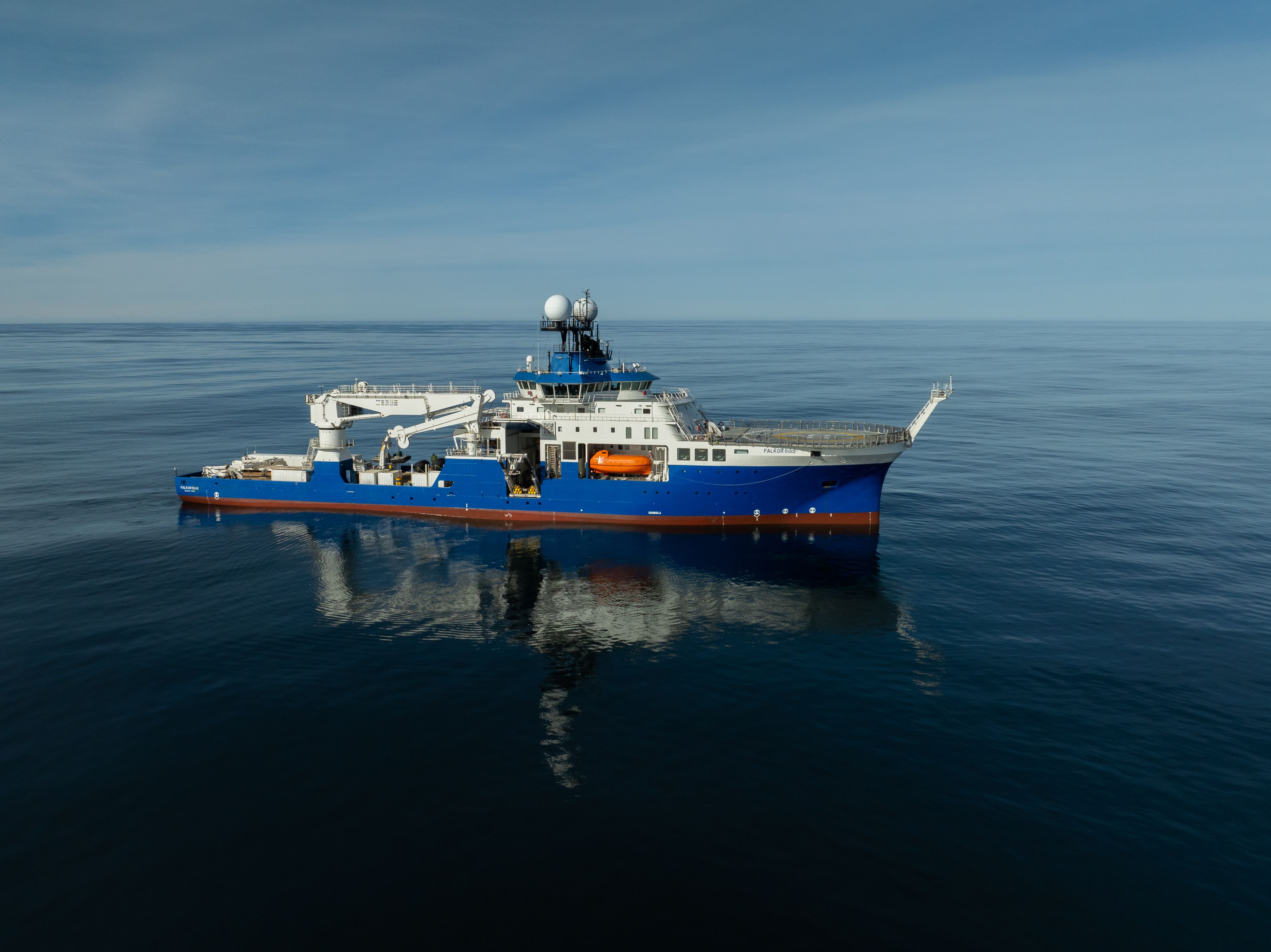 Wide aerial view of the research vessel Falkor (too) resting on calm, clear blue open ocean waters under a sunny sky.