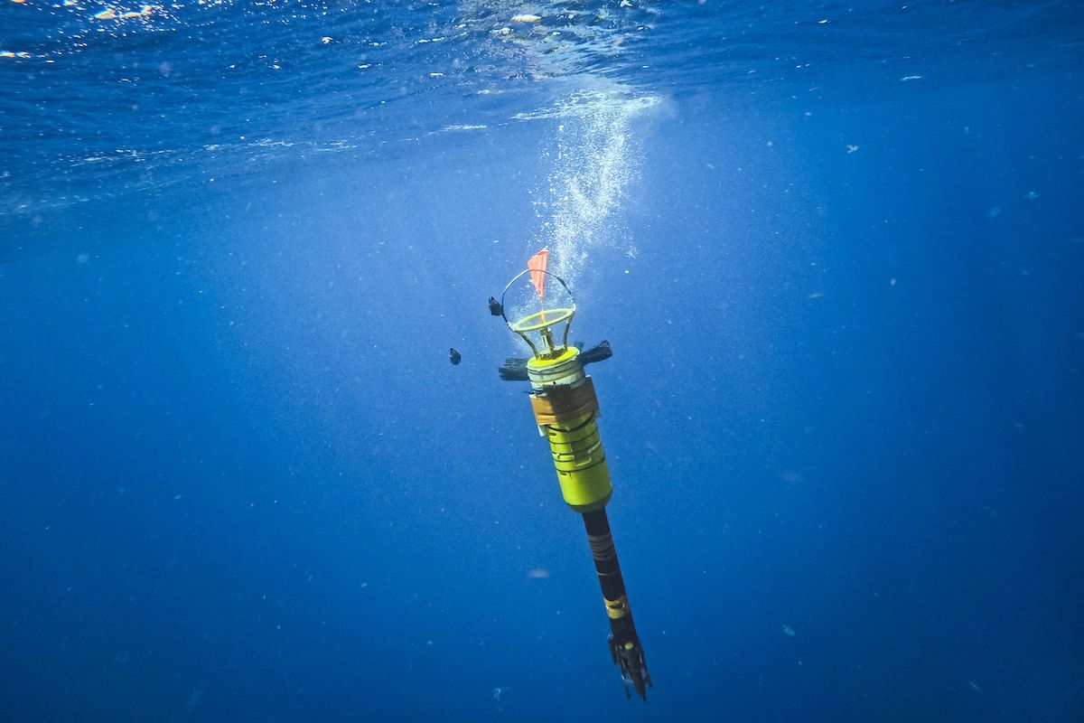A yellow vertical oceanographic profiling instrument (VMP-5500) descending through clear blue water just below the surface, trailing a plume of small air bubbles.