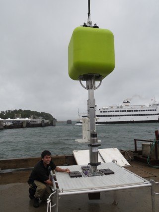 A close up of marine technican James Cooper with the new lander shows the versatile mechanical platform, square metal frame with a perforated top, and tall mast in the middle with the lightweight flotation.