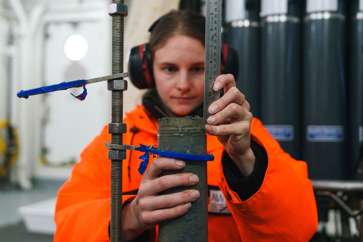 Researcher in an orange jacket measuring a sediment core sample with a metal ruler aboard a vessel.