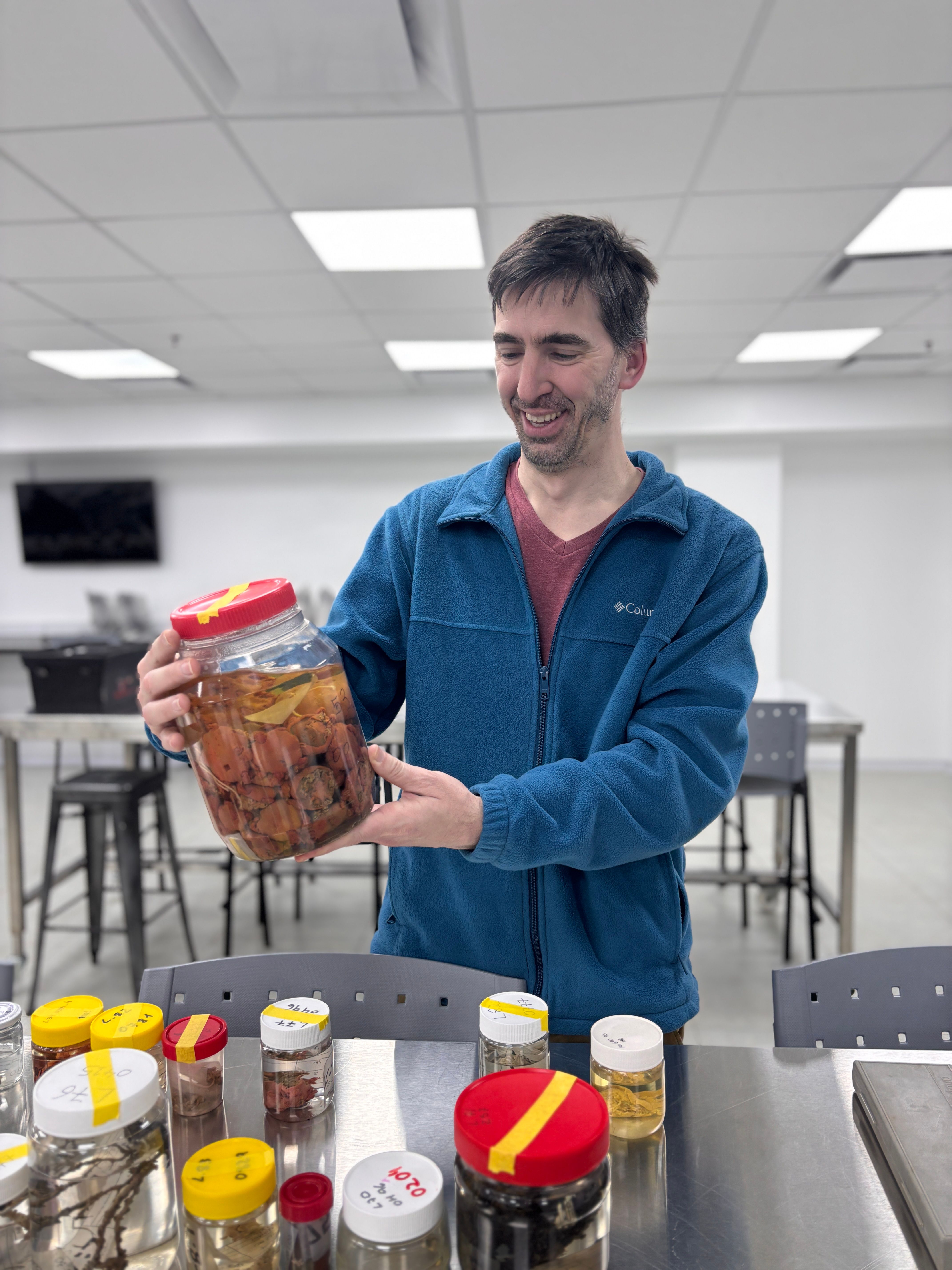 Dr. Daniel Lauretta smiling as he holds up a large glass jar of preserved deep-sea specimens during the Species Discovery Workshop, surrounded by other sample jars on a lab table.