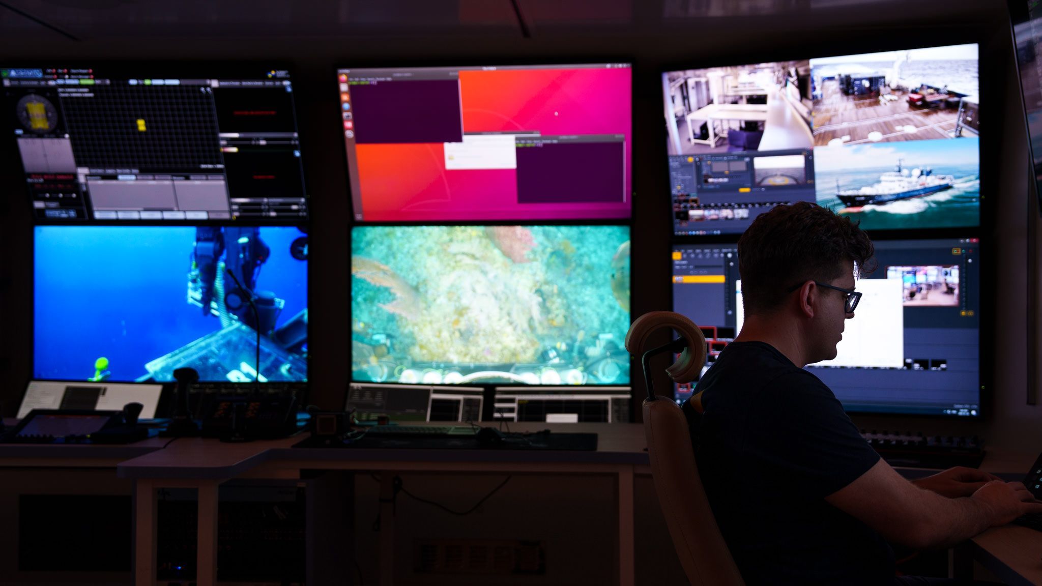 Multimedia technician works on the streaming systems with the mission control monitors in the background. 