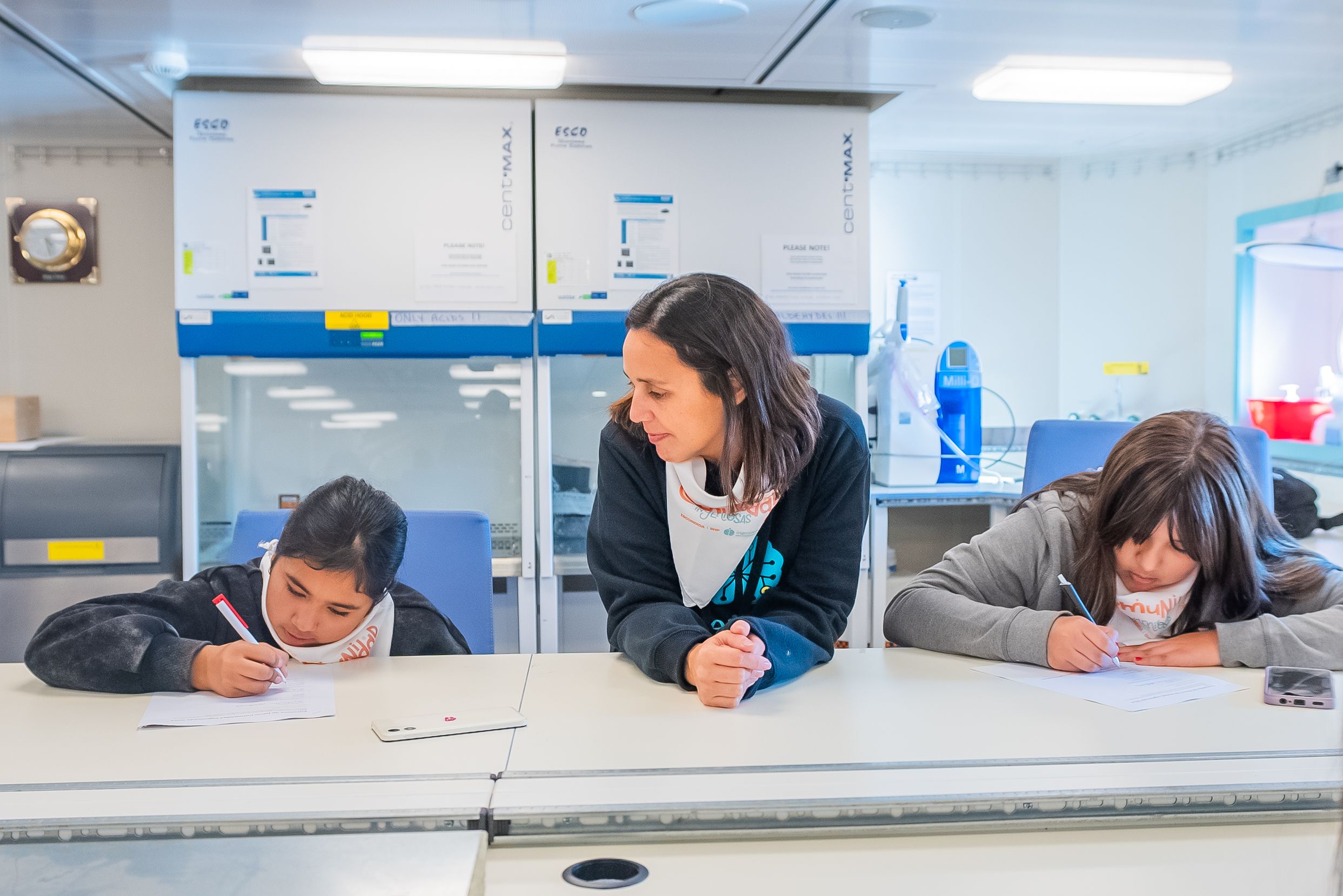A woman with brown hair is sitting with two children who are drawing. She is looking at the one to her left.