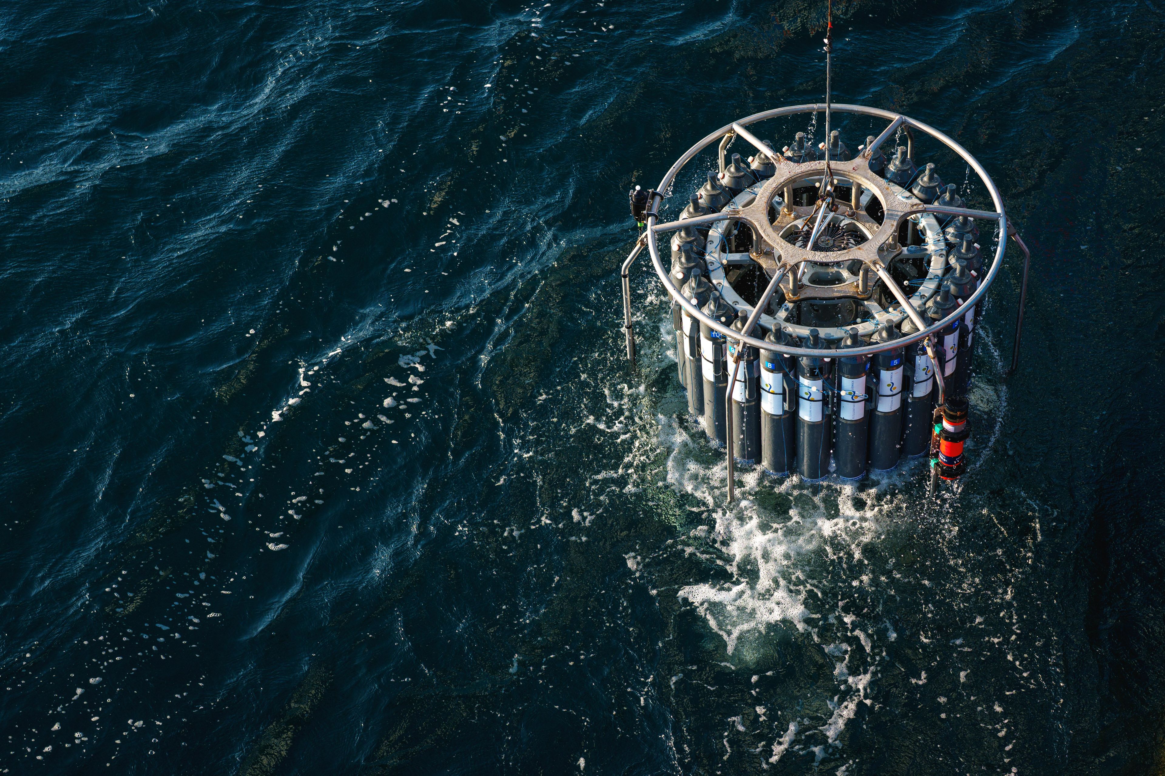 A conductivity, temperature, depth profiler with a rosette of bottles is lifted out of the ocean by a wire.