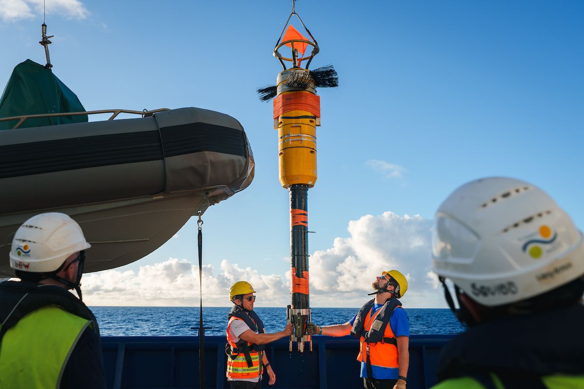 Crew members in hard hats and safety vests guiding the recovery of a tall, yellow VMP-5500 instrument suspended by a crane on the deck of Falkor (too).