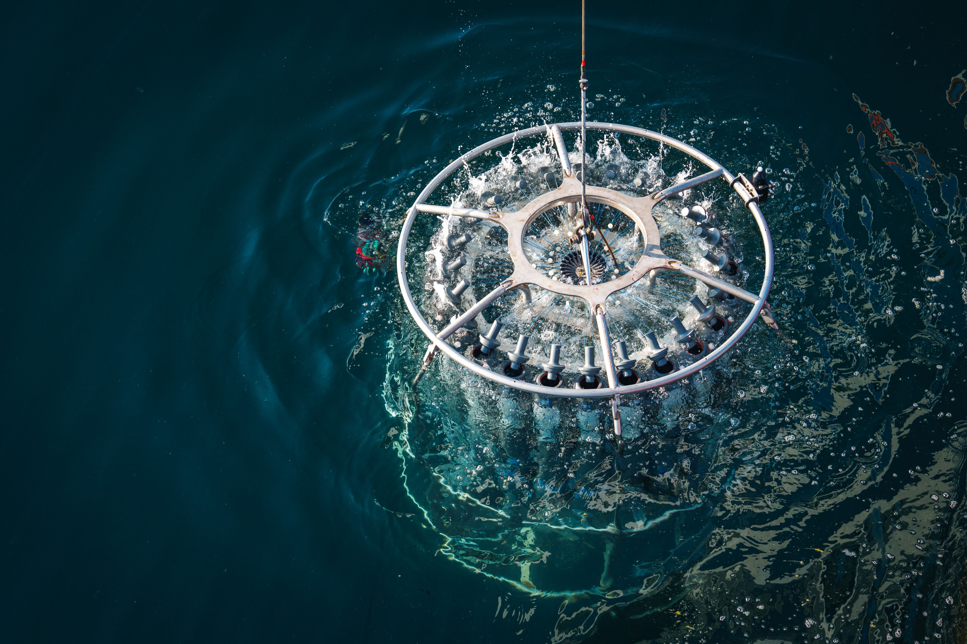 An overhead view of a circular metal CTD rosette being lowered into or lifted out of deep blue ocean water, creating white splashes and ripples around its frame.