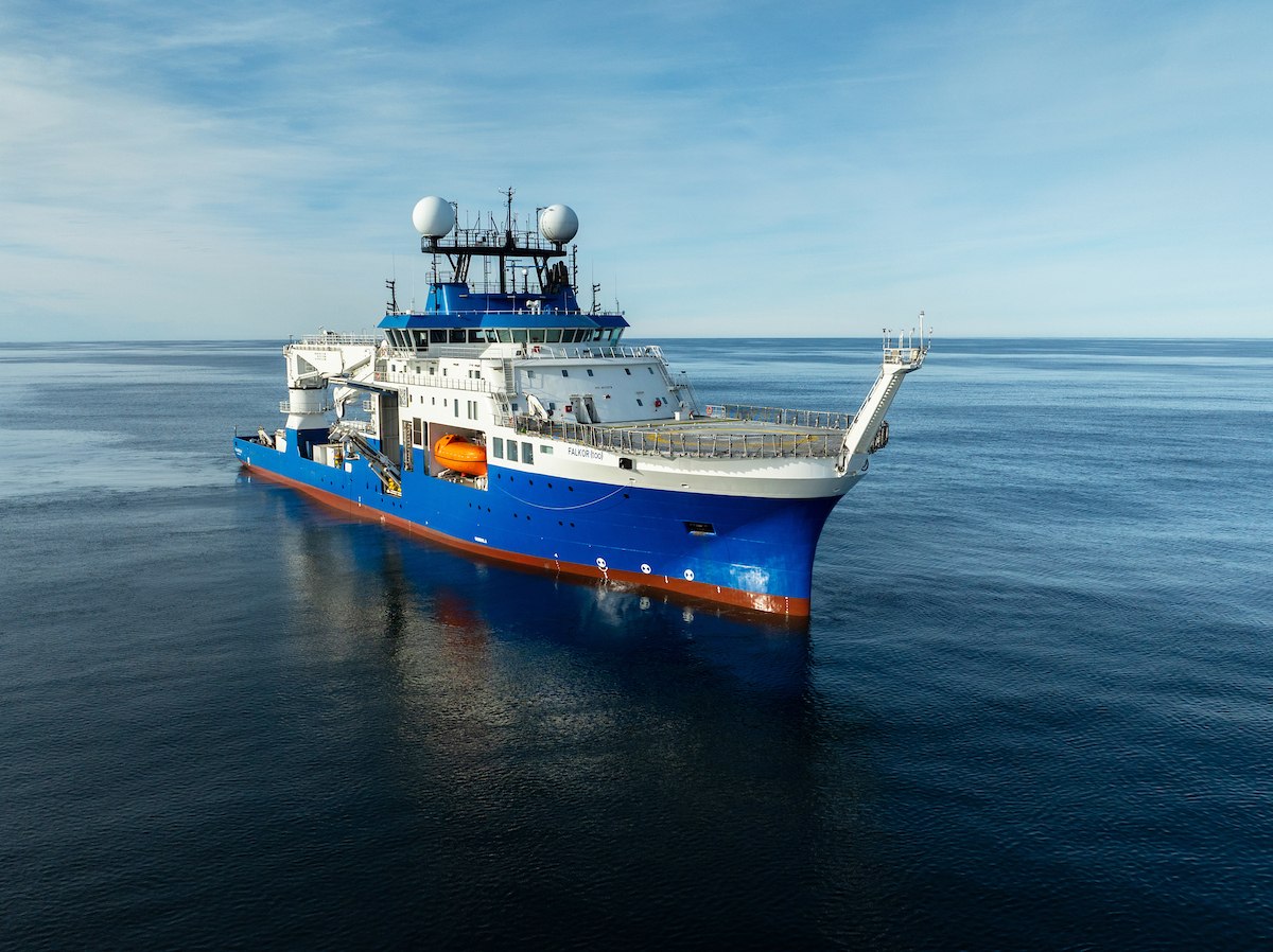 Research vessel Falkor (too) with its blue hull, white superstructure, and radar domes anchored on calm, deep blue ocean waters under a clear sky.
