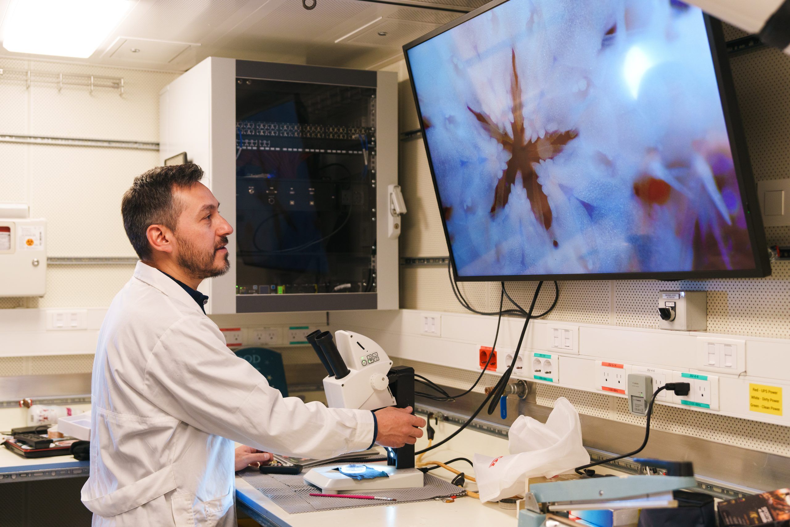 A scientist works in a lob, looking at a screen attached to a microscope.