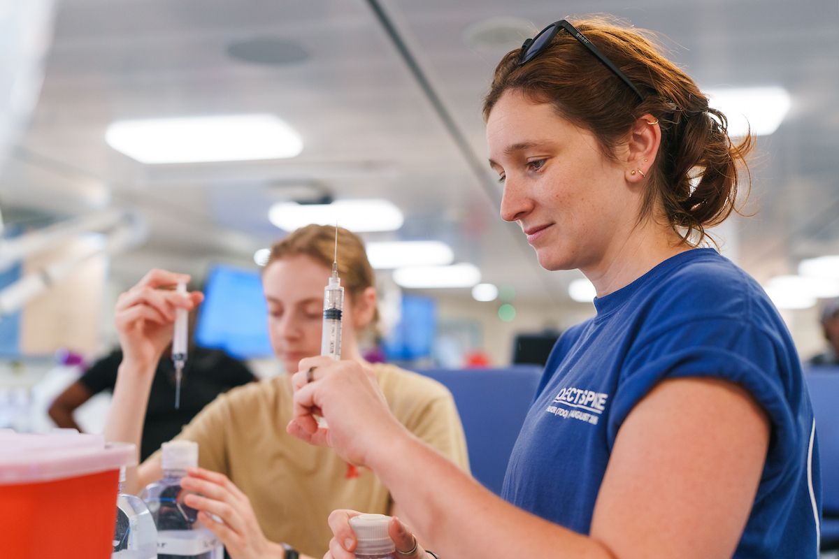 A researcher using a syringe to draw water samples from a bottle during lab work aboard the vessel, with a colleague working in the background.