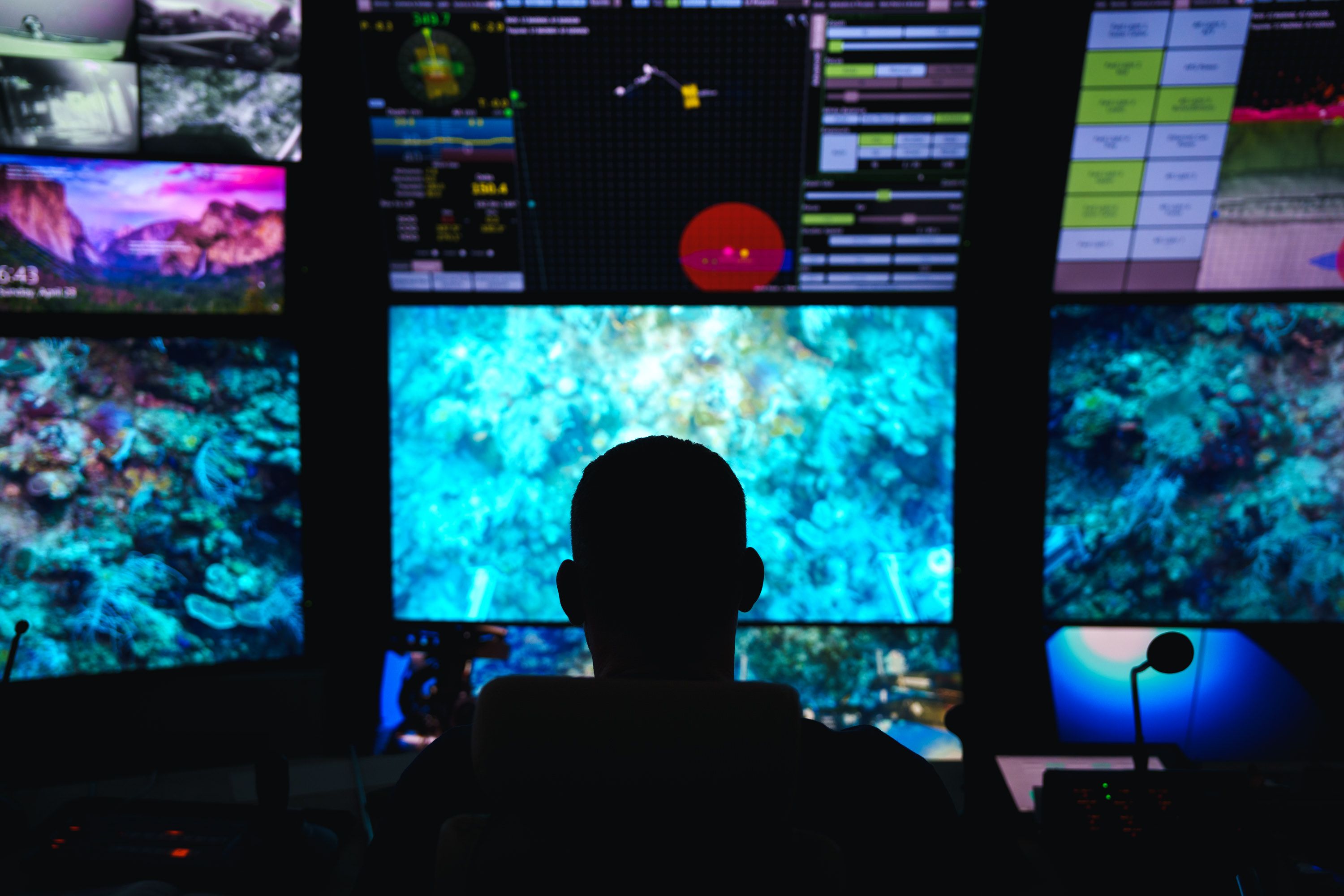 Silhouette of a beloved ROV pilot with several screens infront of him showing live imagery from the Remotely Operated Vehicle he is piloting.    
