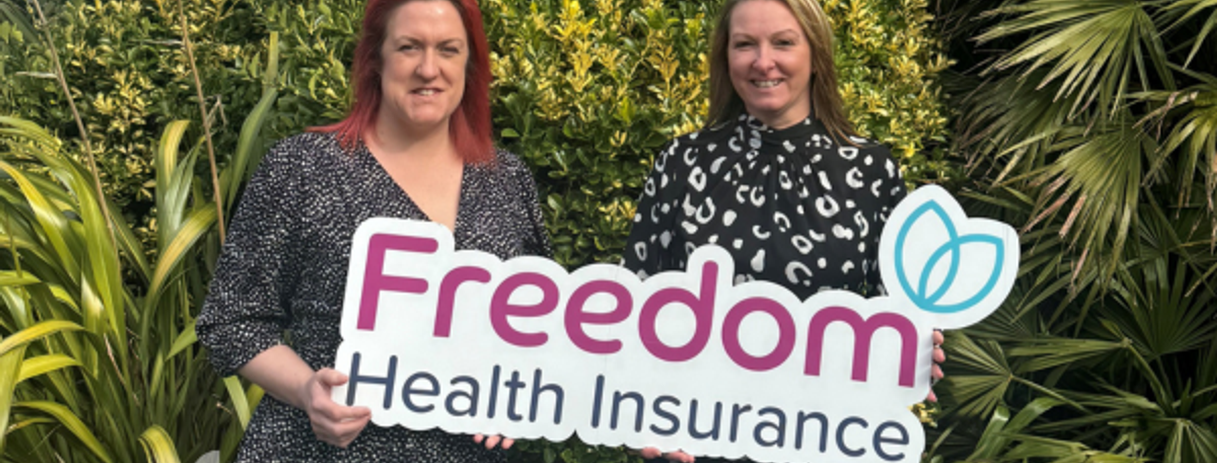 Two women holding a 'Freedom Health Insurance' sign outdoors, smiling amid green foliage.