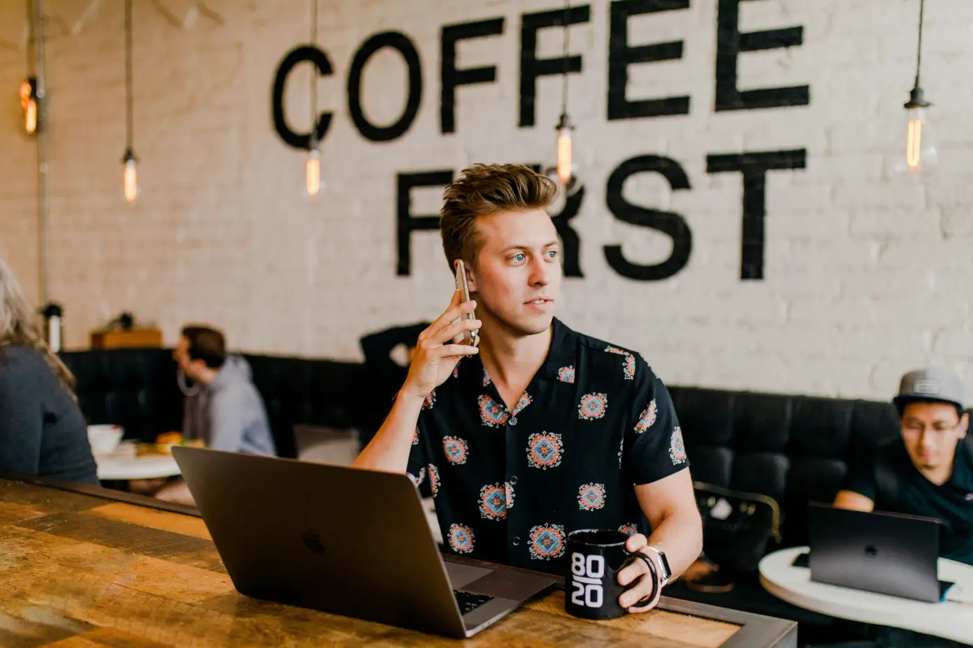 Mladý muž sedí v kavárně s notebookem, udržuje oční kontakt mimo kameru a drží šálek kávy, zatímco nápis "Coffee First" je viditelný na pozadí.