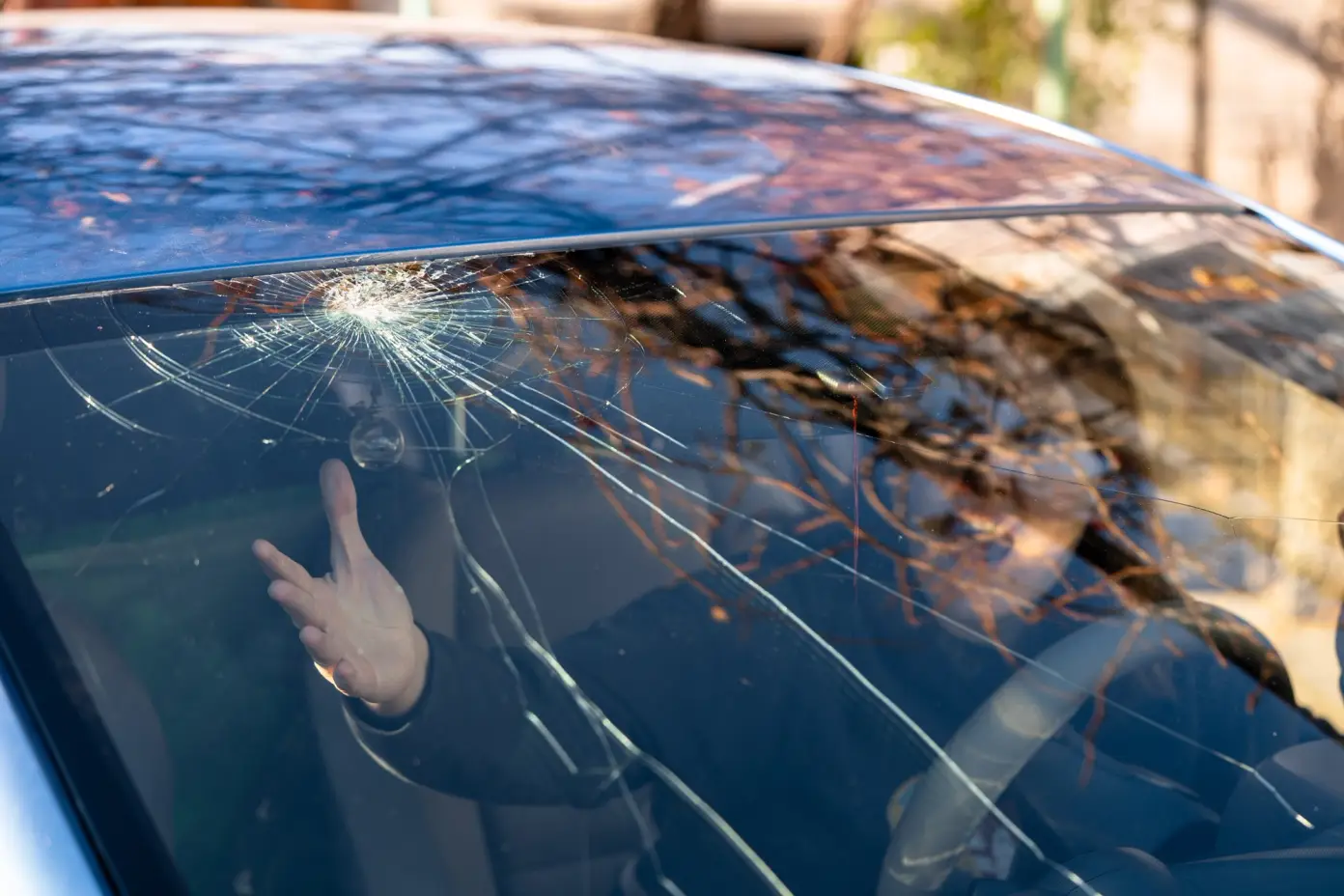 Pohled na prasklé čelní sklo automobilu, z pohledu zevnitř, s osobou uvnitř ukazující rukou na prasklinu.