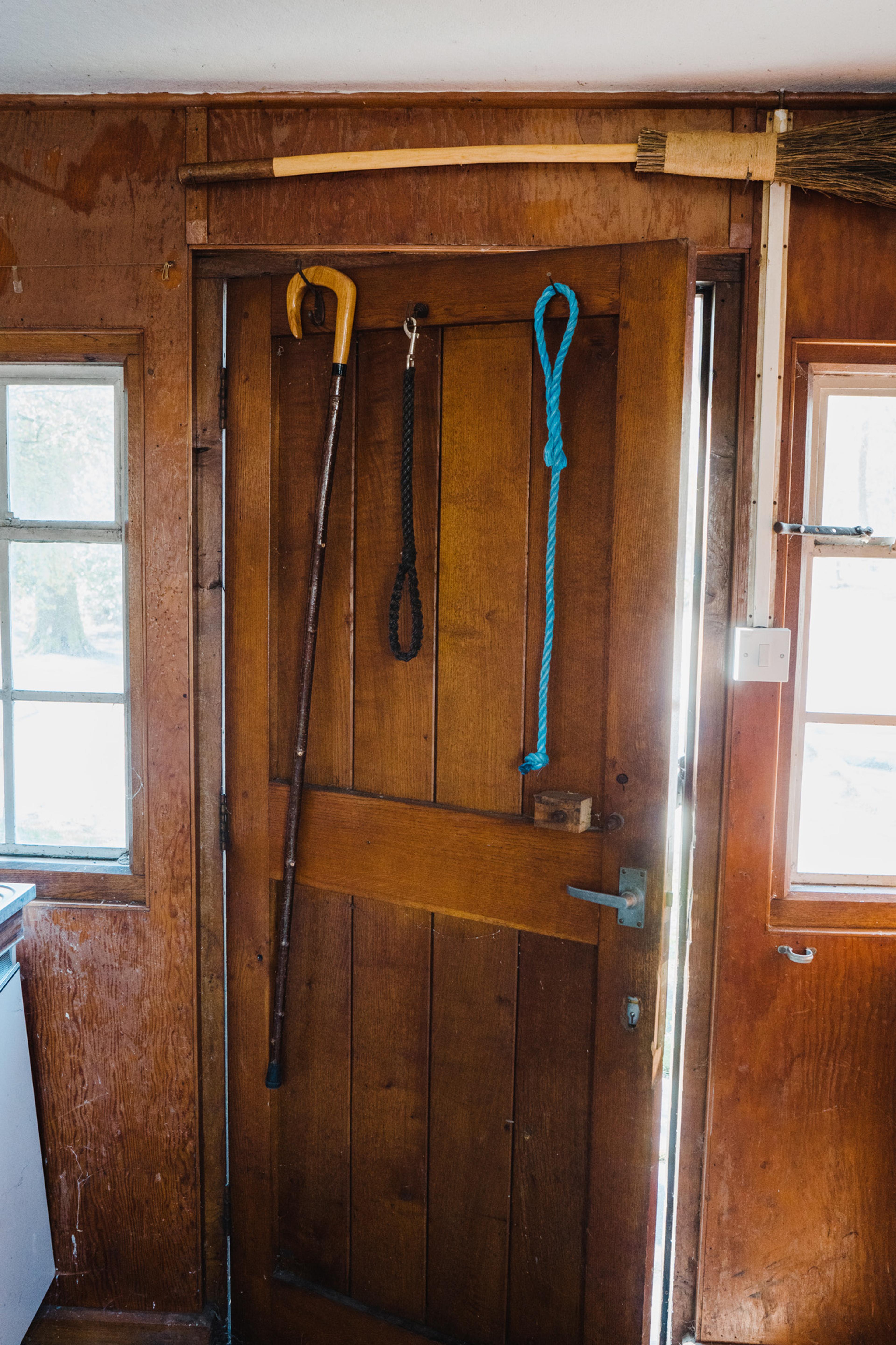 A wooden door with a cane and two dog leads hanging on it, and a broom hung on the wall above the door