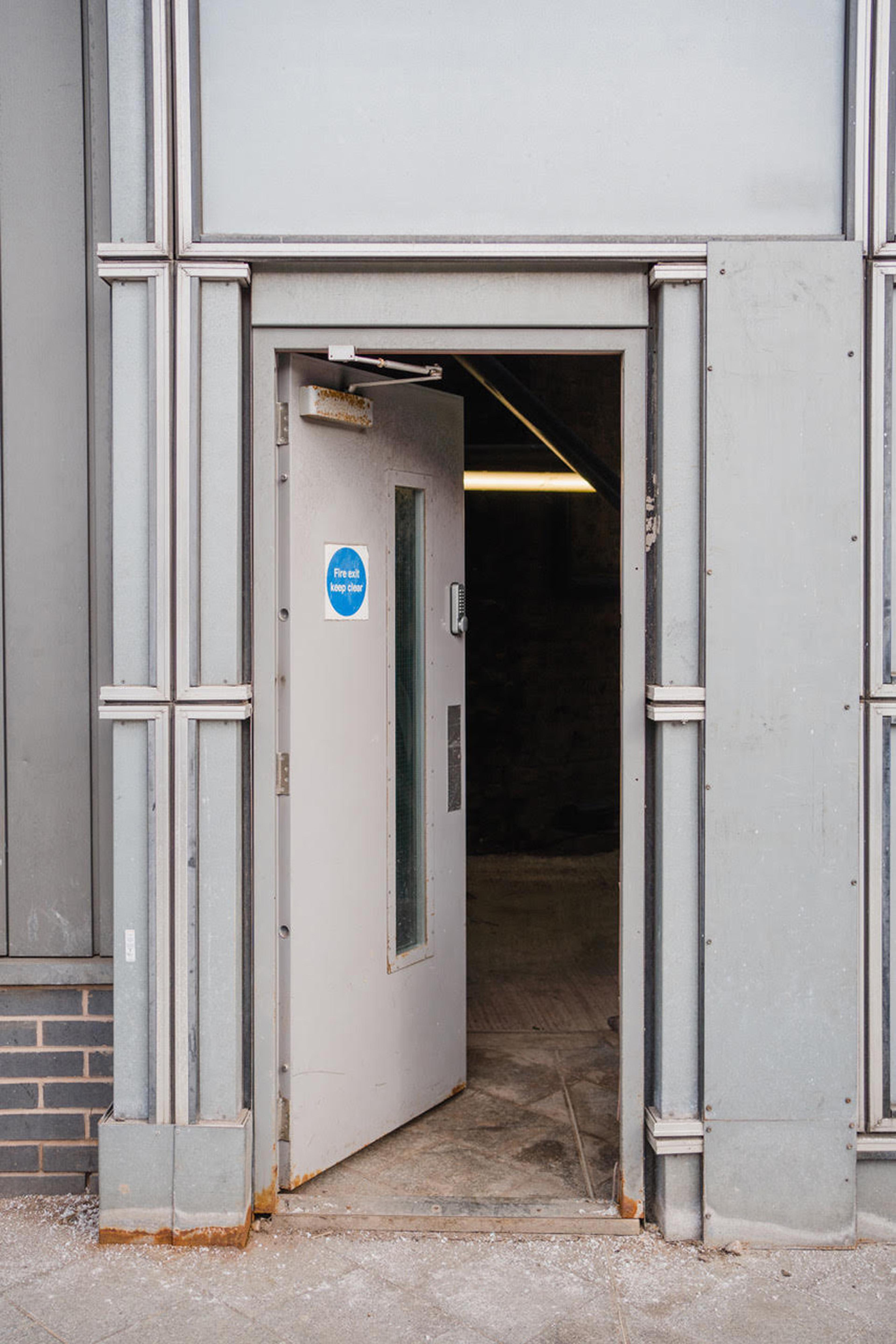 A grey door with a blue fire exit sticker is open to a dark space