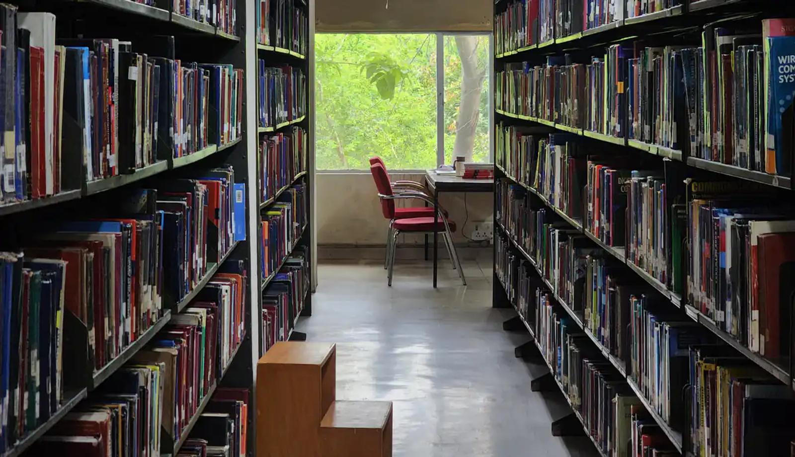 Kütüphanede iki uzun kitap rafı arasında pencere önündeki kırmızı sandalye ve masa