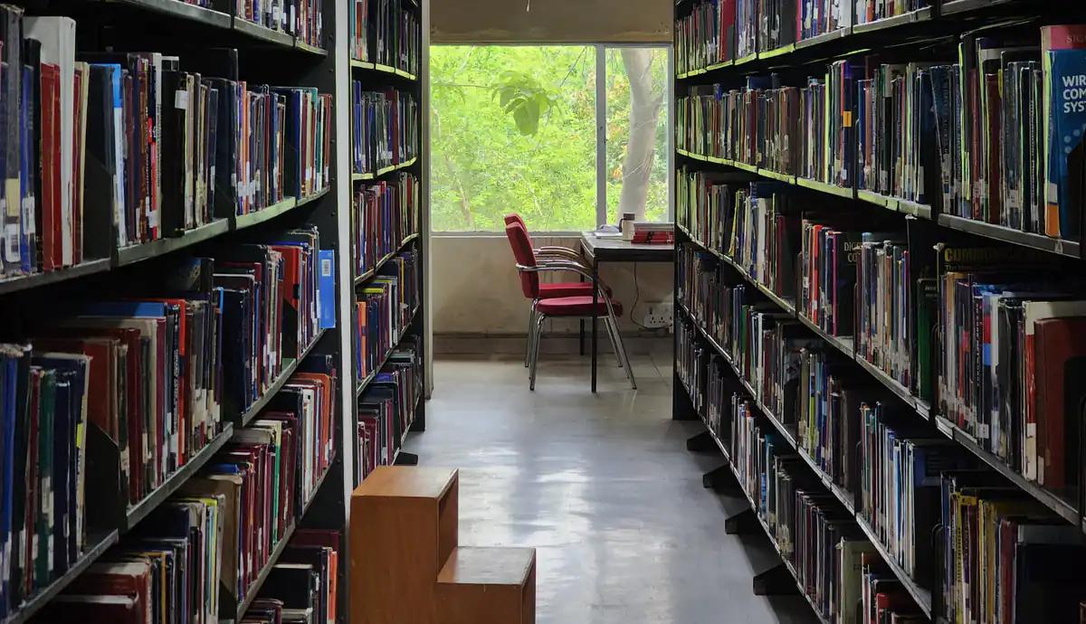 Kütüphanede iki uzun kitap rafı arasında pencere önündeki kırmızı sandalye ve masa