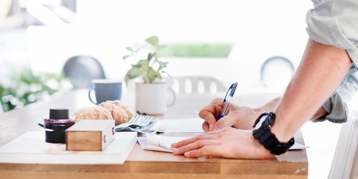 Ahşap bir masada ayakta durarak kalemle bir belgeye not yazan bir kişinin elleri