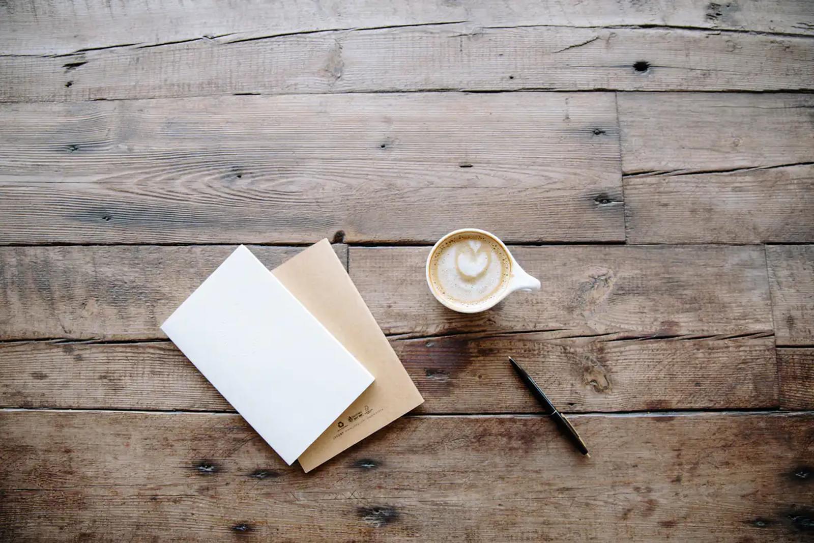 Ahşap masa üzerinde iki defter, bir fincan kahve ve bir kalem
