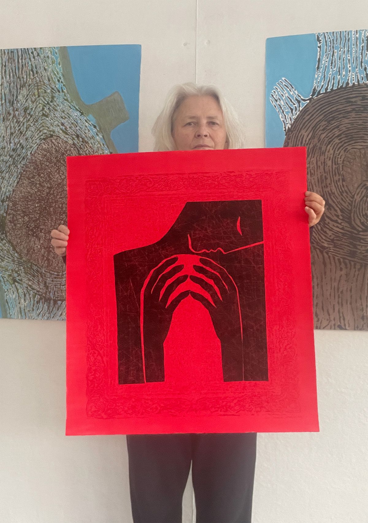 Jolanta holding her red-and-black linocut Inner Gate in her studio, with two large semi-abstract paintings on the wall behind her.