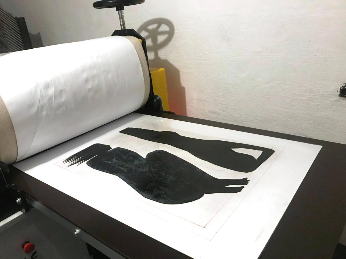 A matrix from Jolanta Johnsson's Human series lying on the bed of the etching press in her studio, the roller and blanket drawn back, two black cut figures on white paper.