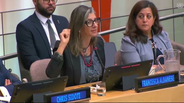A woman sitting on a panel speaks into the mic at a meeting and gestures with her hand. 