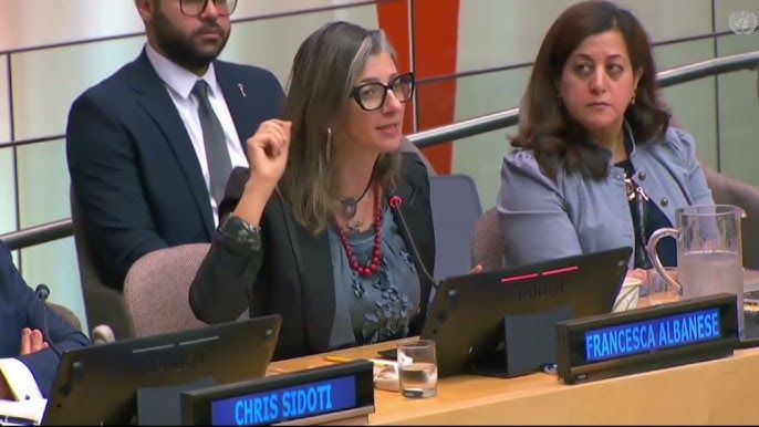 A woman sitting on a panel speaks into the mic at a meeting and gestures with her hand.