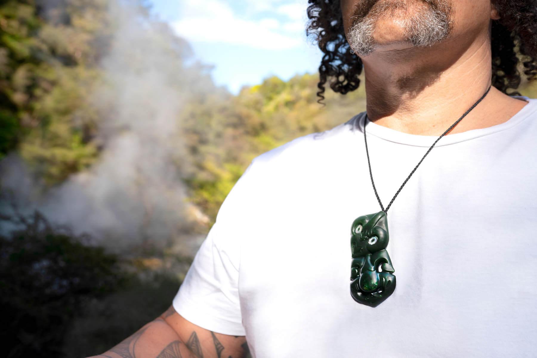 Steam rises in geothermal setting behind man wearing New Zealand Pounamu Hei Tiki