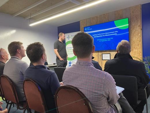 The picture shows the audience at the Build Test Solutions Open Day 2023 listening to Richard Jack speaking about building test solutions and best practices. There is a presentation running on the big screen while engineers are sat watching and taking notes.