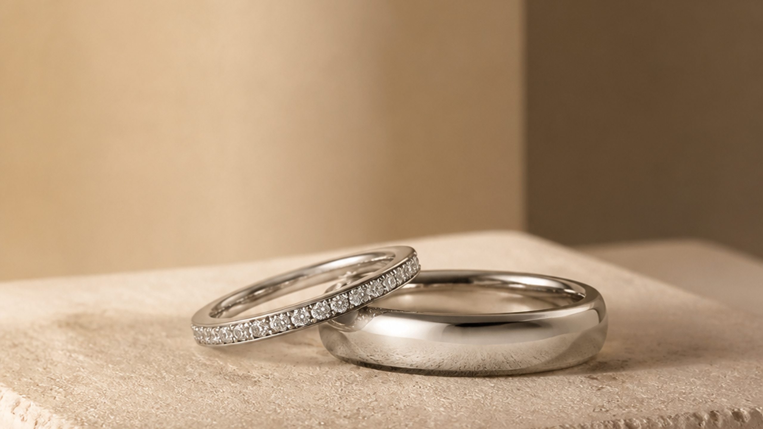 Editorial photograph of a polished wedding band and a diamond-set band on pale stone