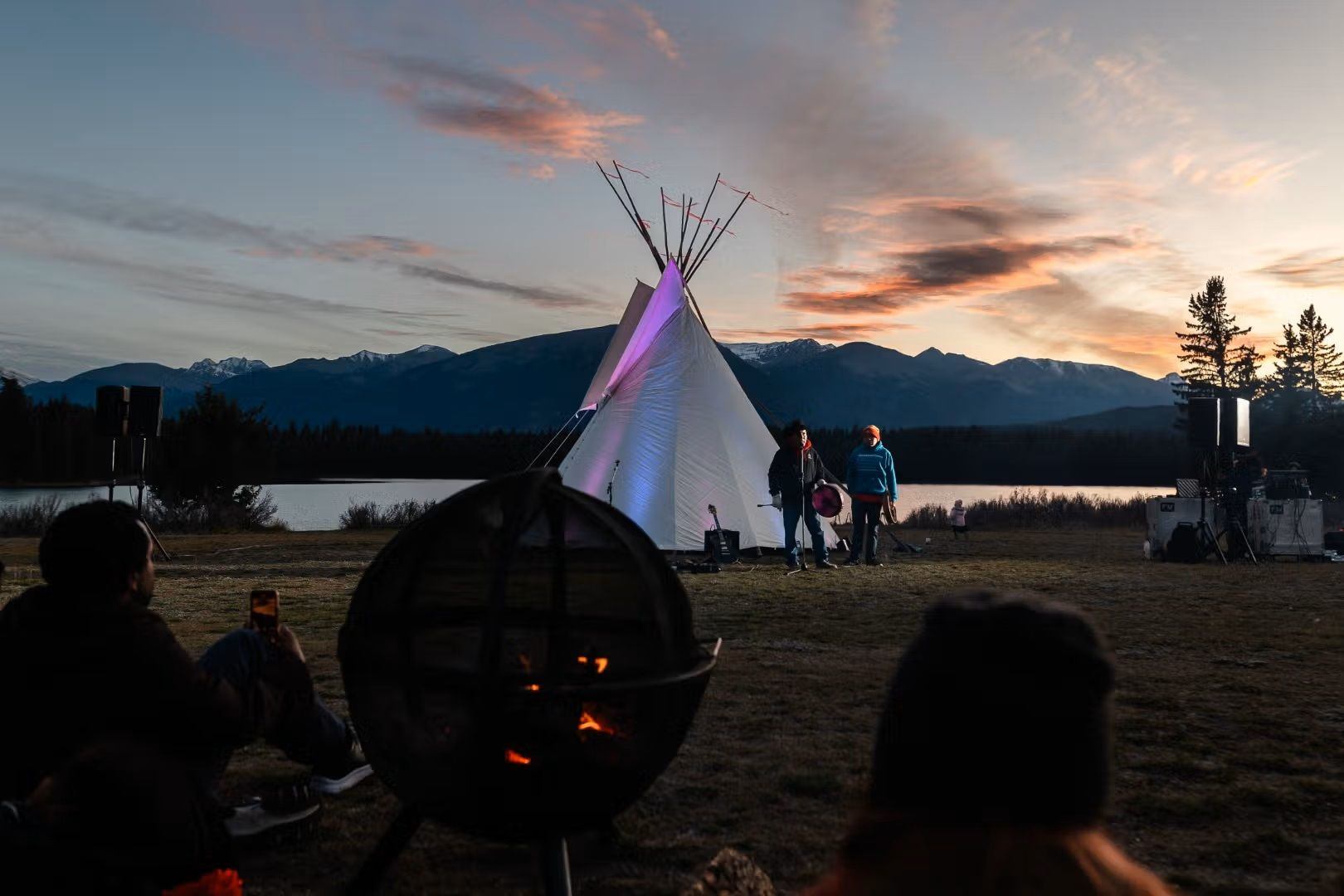 A fire burns in an enclosed firepit with a teepee and alpine lake in the background.