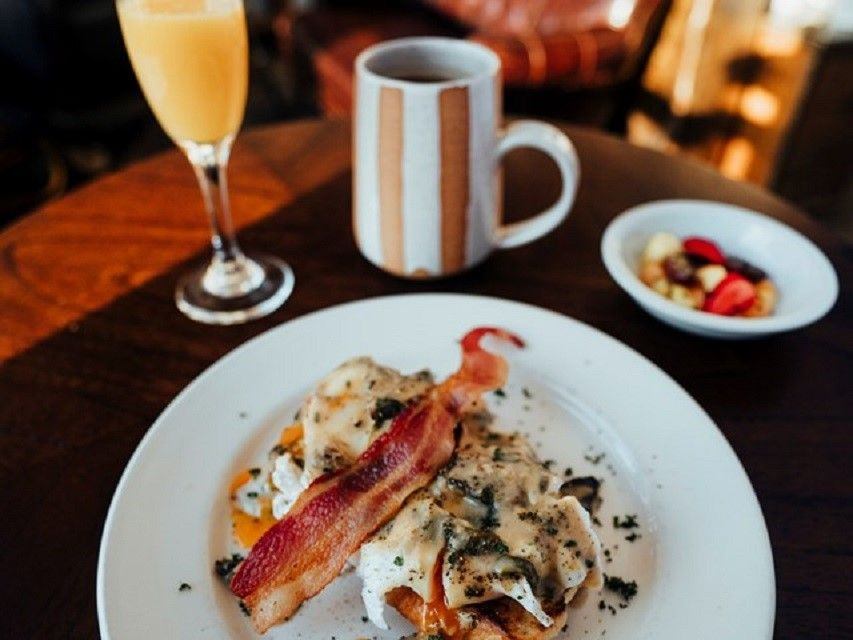 A strip of bacon sits atop eggs with a coffee mug and glass of orange juice nearby.