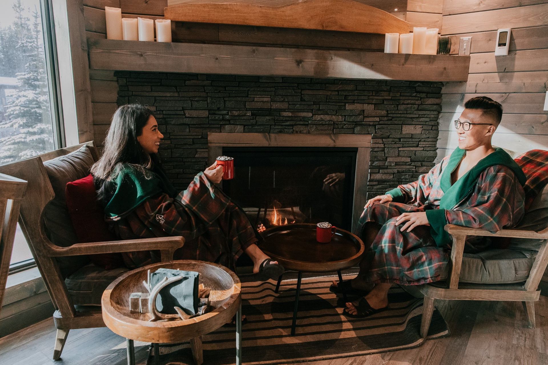 A couple dressed in plaid robes sits near a fireplace on a winter day.