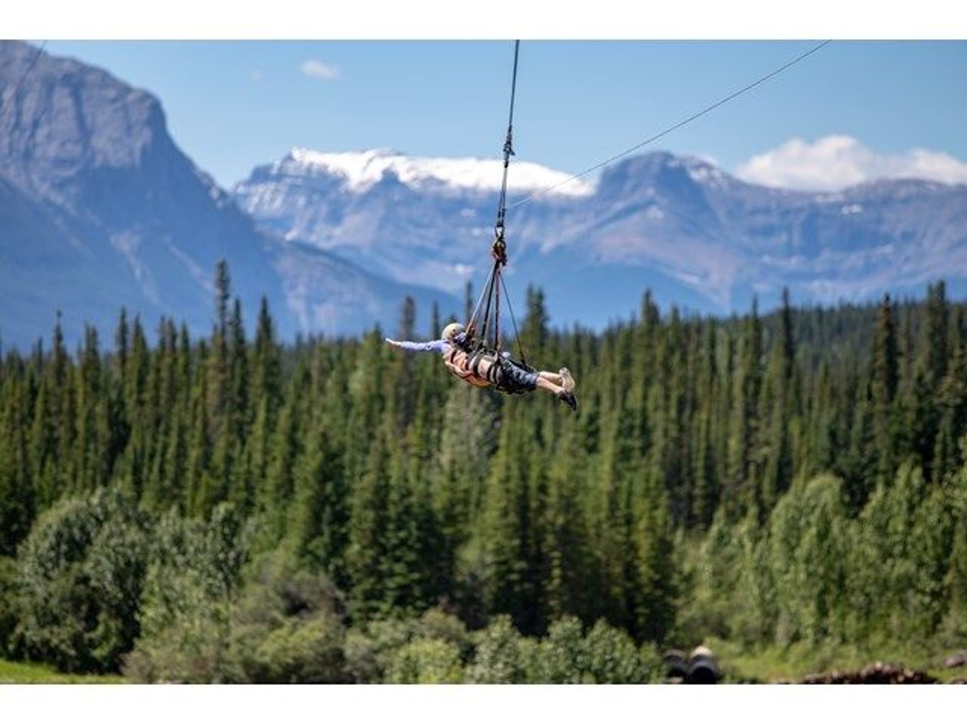A person soars on a zipline above Alberta forests with the Canadian Rockies in the background.