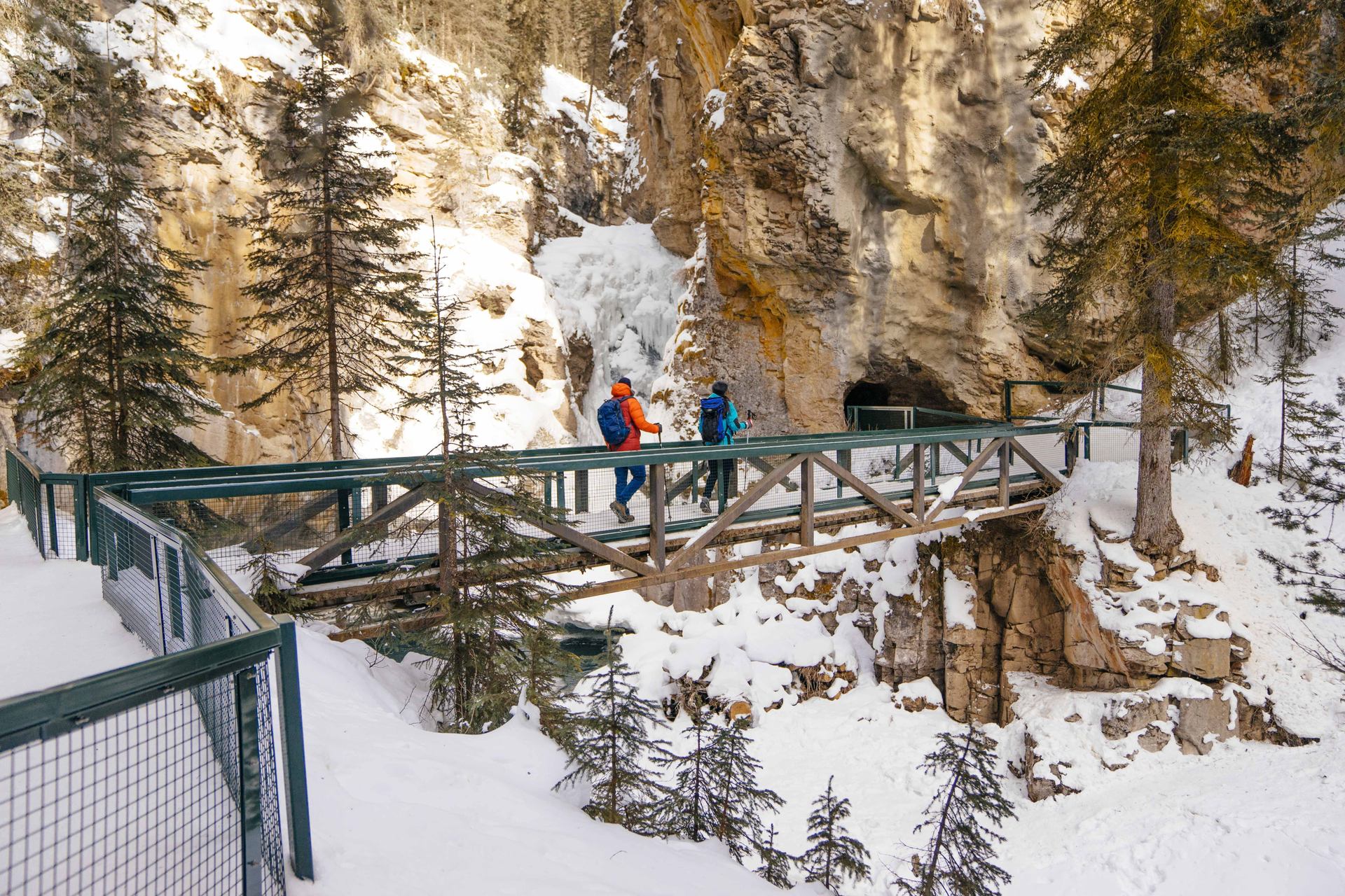 A couple winter hiking over a bridge heading toward a cave opening to the lower falls at Johnston Canyon.