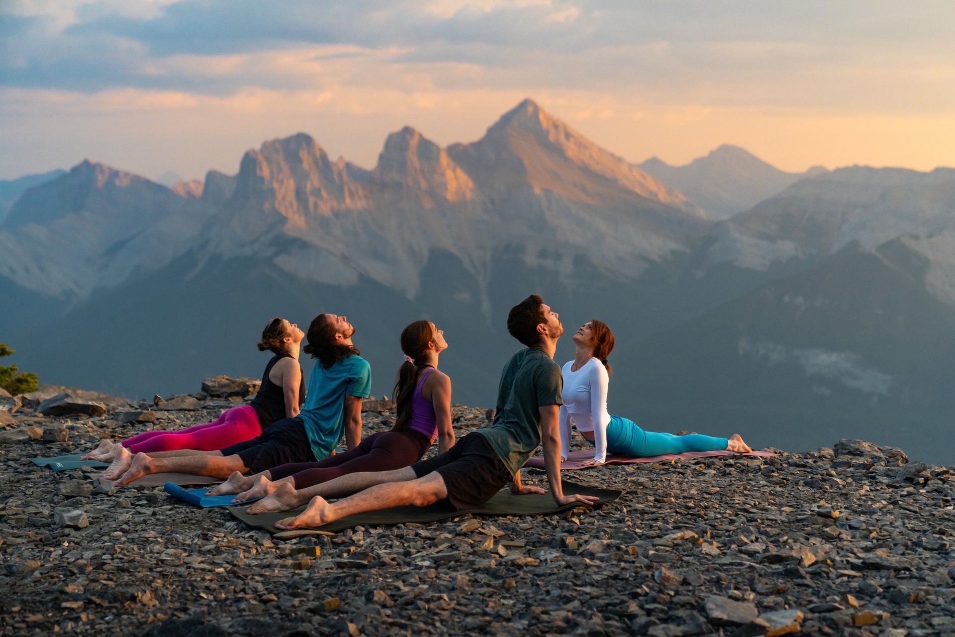 Five people do yoga on top of Mount Lady MacDonald with the Three Sisters looming large in the background.