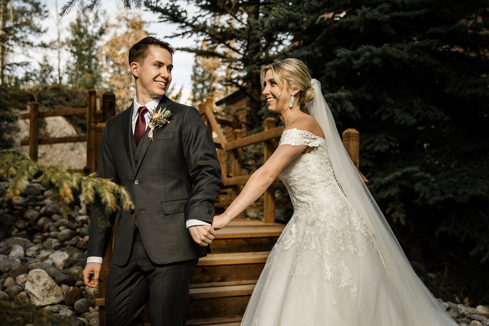 A groom and bride in wedding gown smile while holding hands.