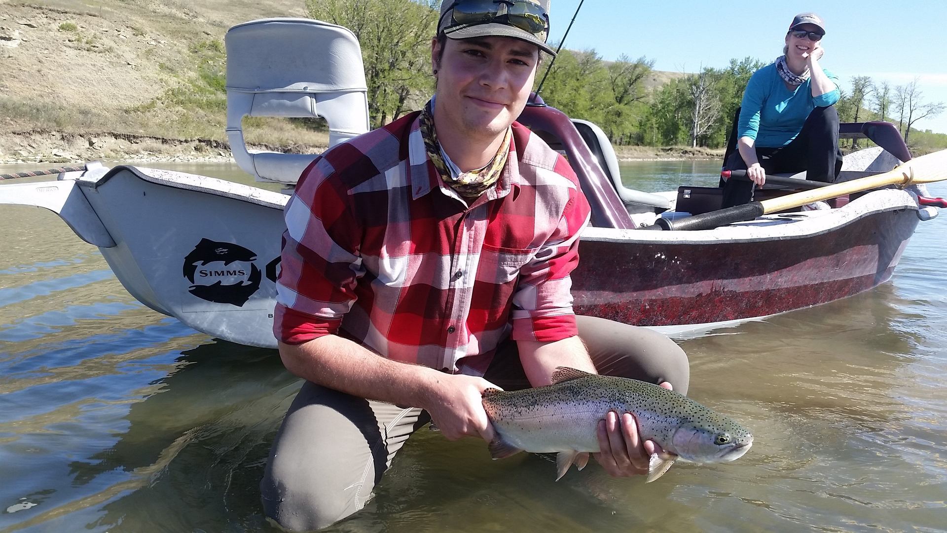 A young man in a plaid shirt squats in the water holding a fish.