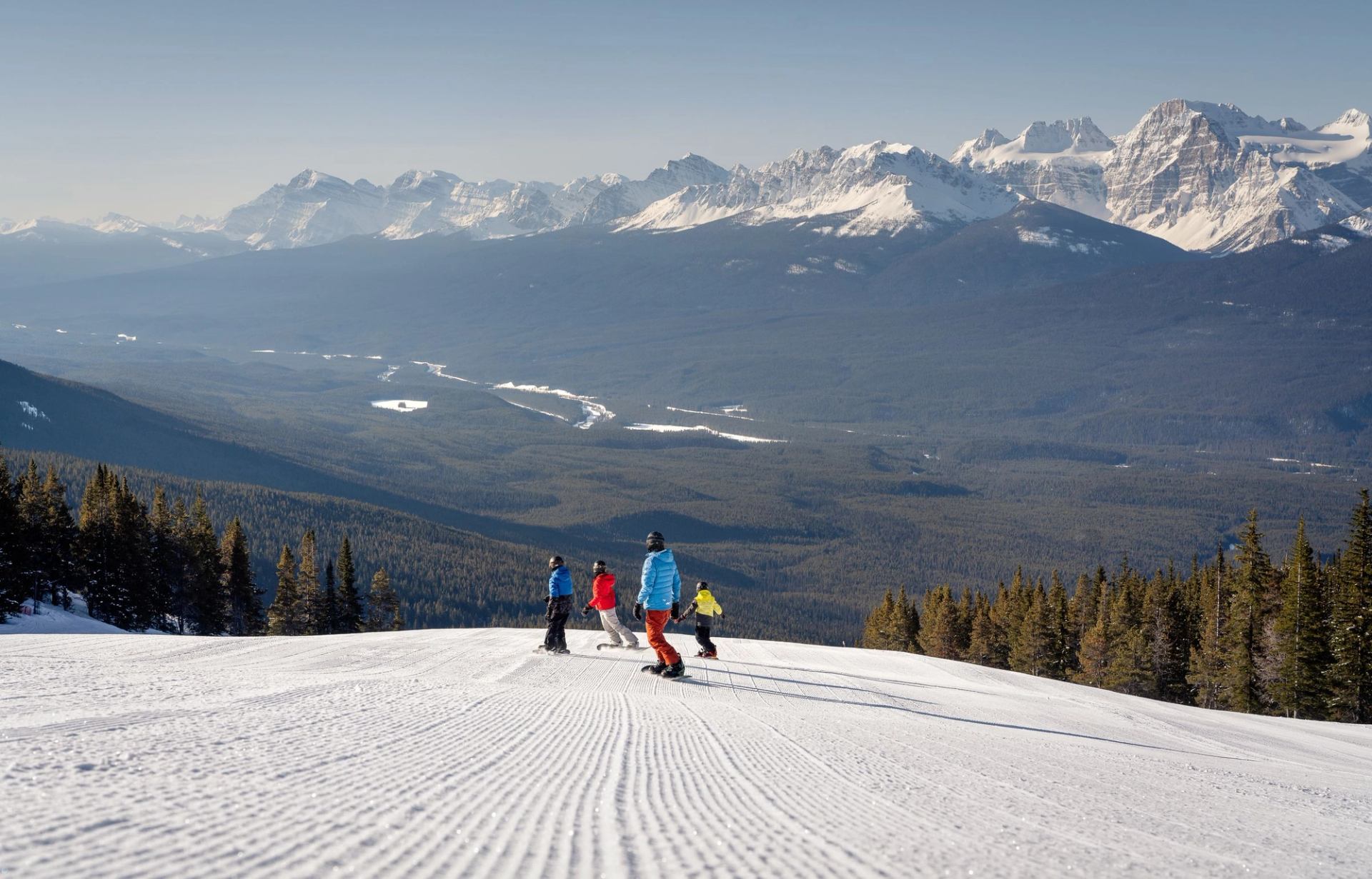 A group of four snowboarders carve on an open groomed run with mountain views while snowboarding at Lake Louise Ski Resort in Banff National Park