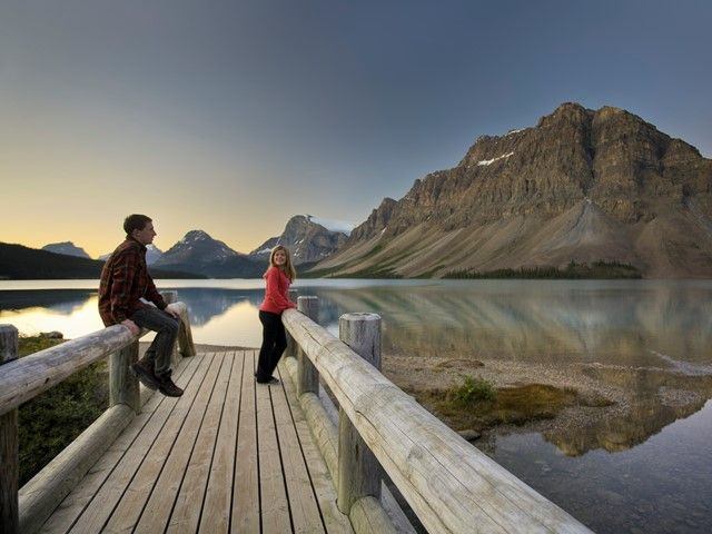 Couple standing on wooden dock with mountains reflecting in the water at Bow Lake in Banff National Park.